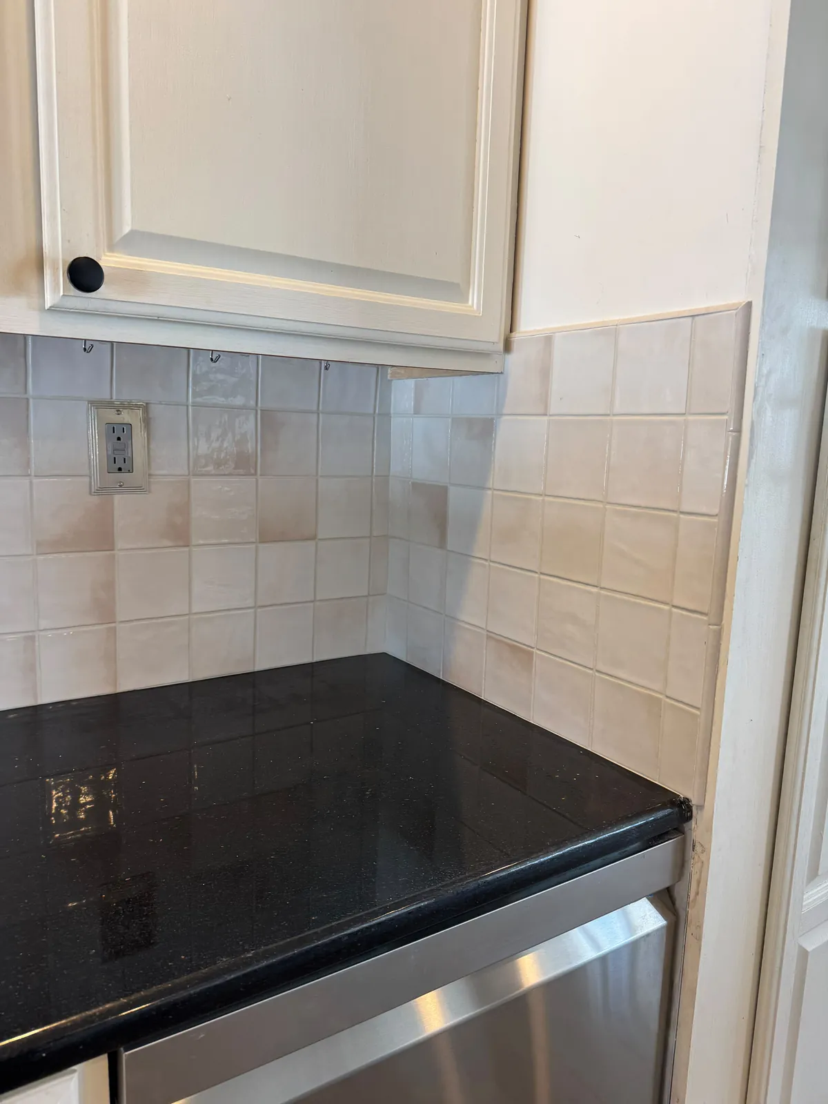 Inside corner where a cream square tile backsplash turns onto the side wall, above a black granite counter with white cabinets overhead