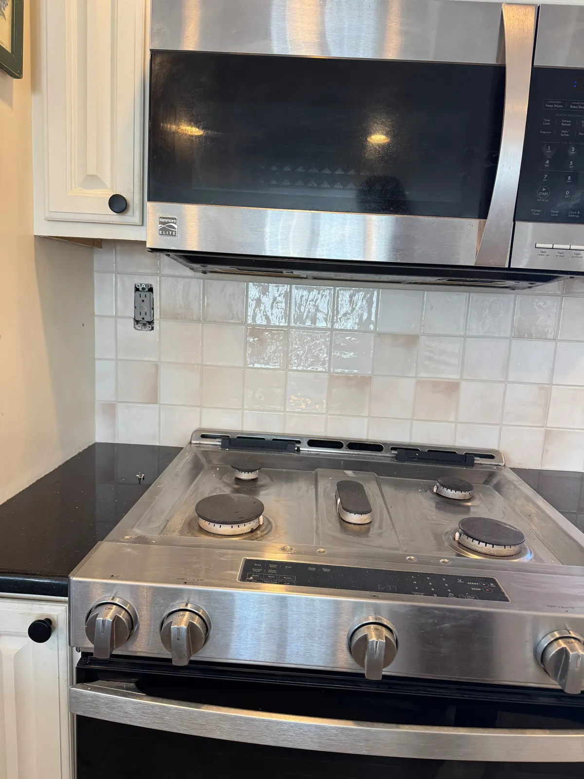 Cream and blush square handmade-look tile backsplash behind a gas range and below a microwave, with black granite counters and white cabinets