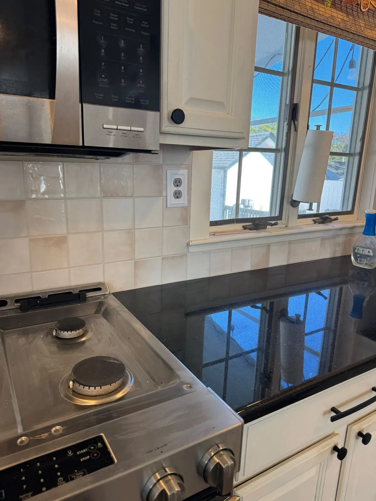 Cream square tile backsplash running from beside a gas range to a kitchen window, with the window reflected in a black granite counter