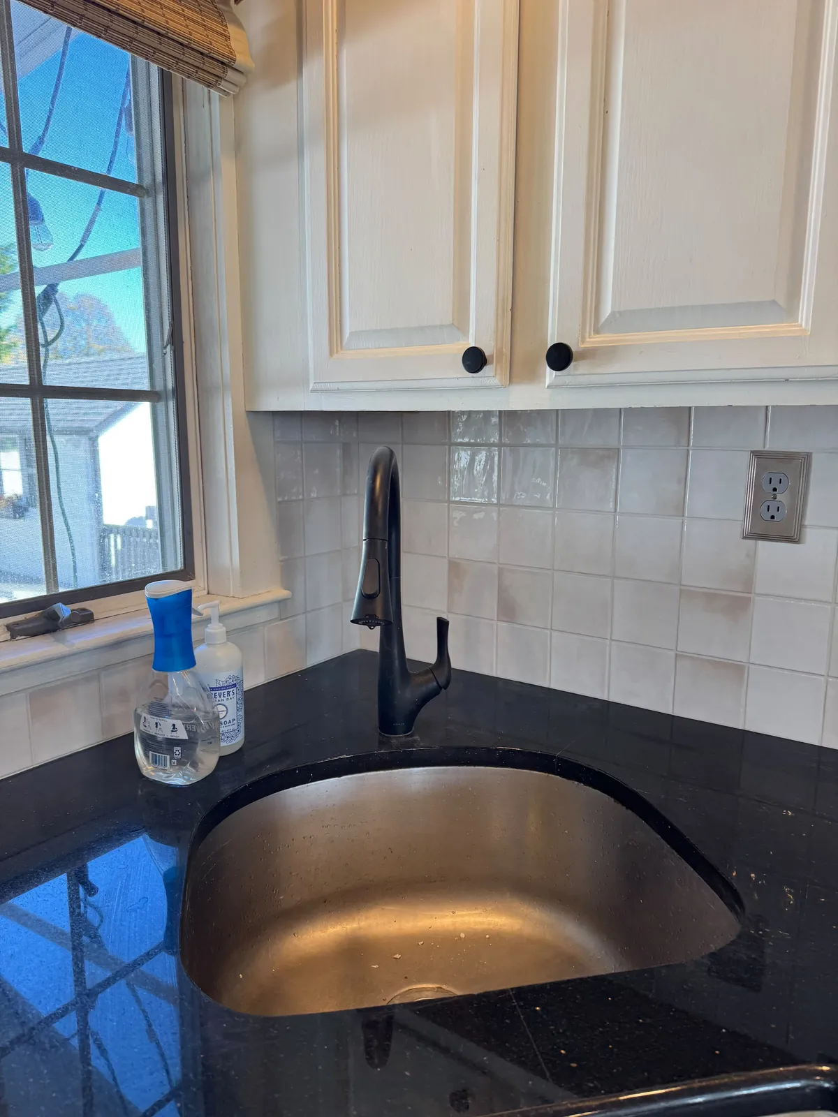 Cream square tile backsplash around a corner sink window, with a black pull-down faucet, an undermount sink and black granite counters