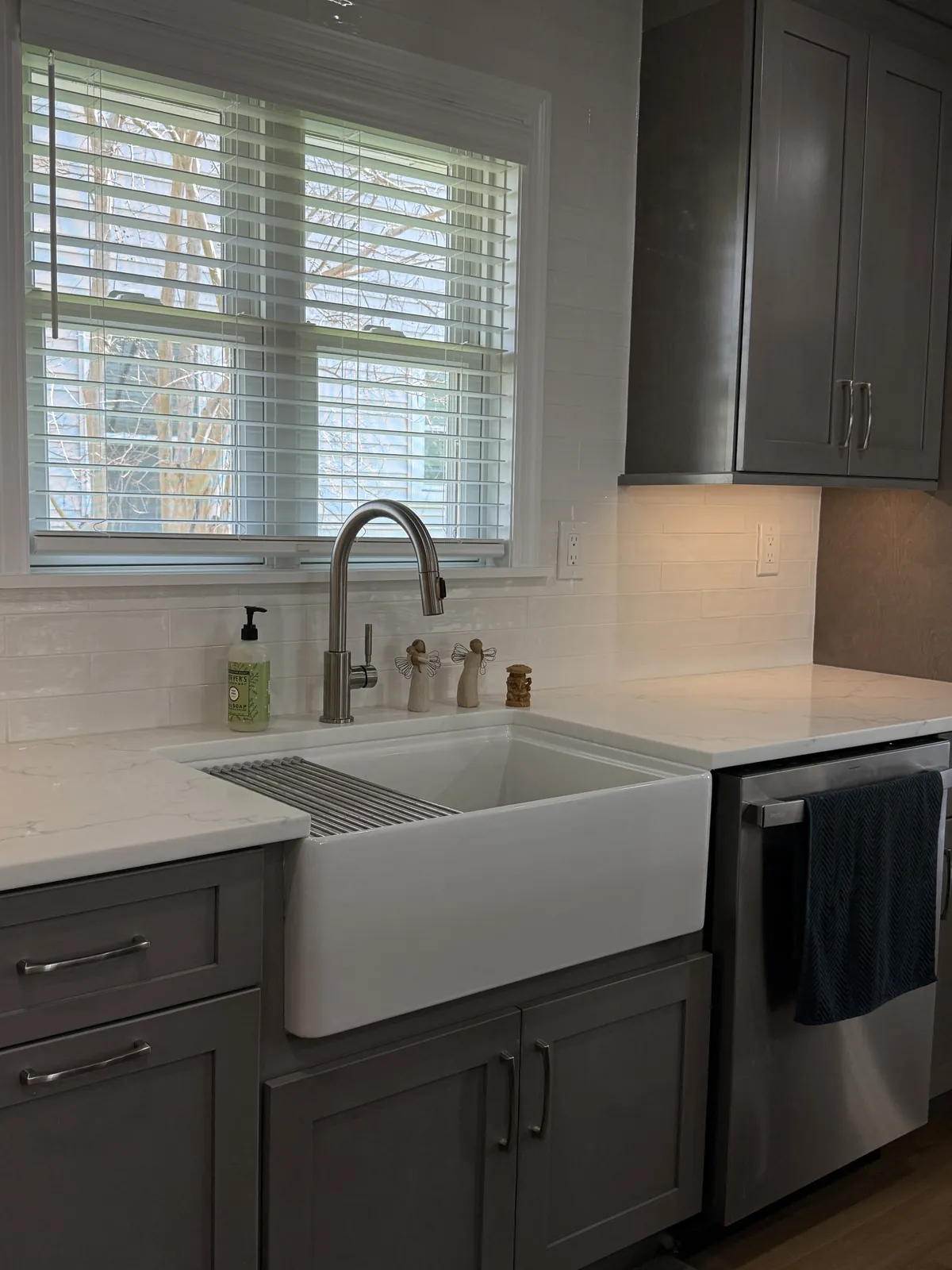 White subway tile backsplash behind a farmhouse sink under a window, with gray cabinets and a stainless dishwasher