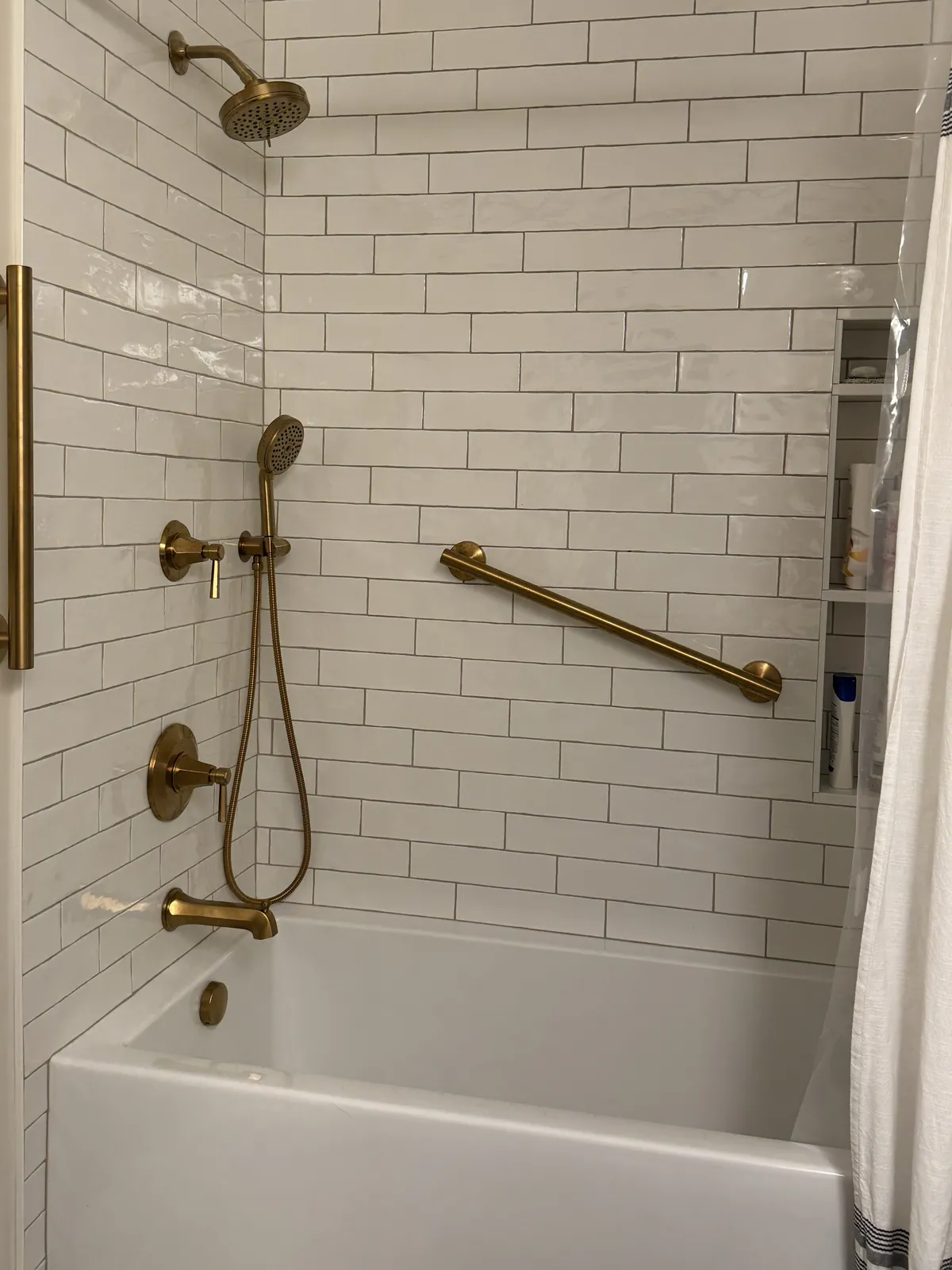 Tub wall tiled in long white subway tile with brushed brass shower head, hand shower, slide bar and grab bars, and a recessed niche