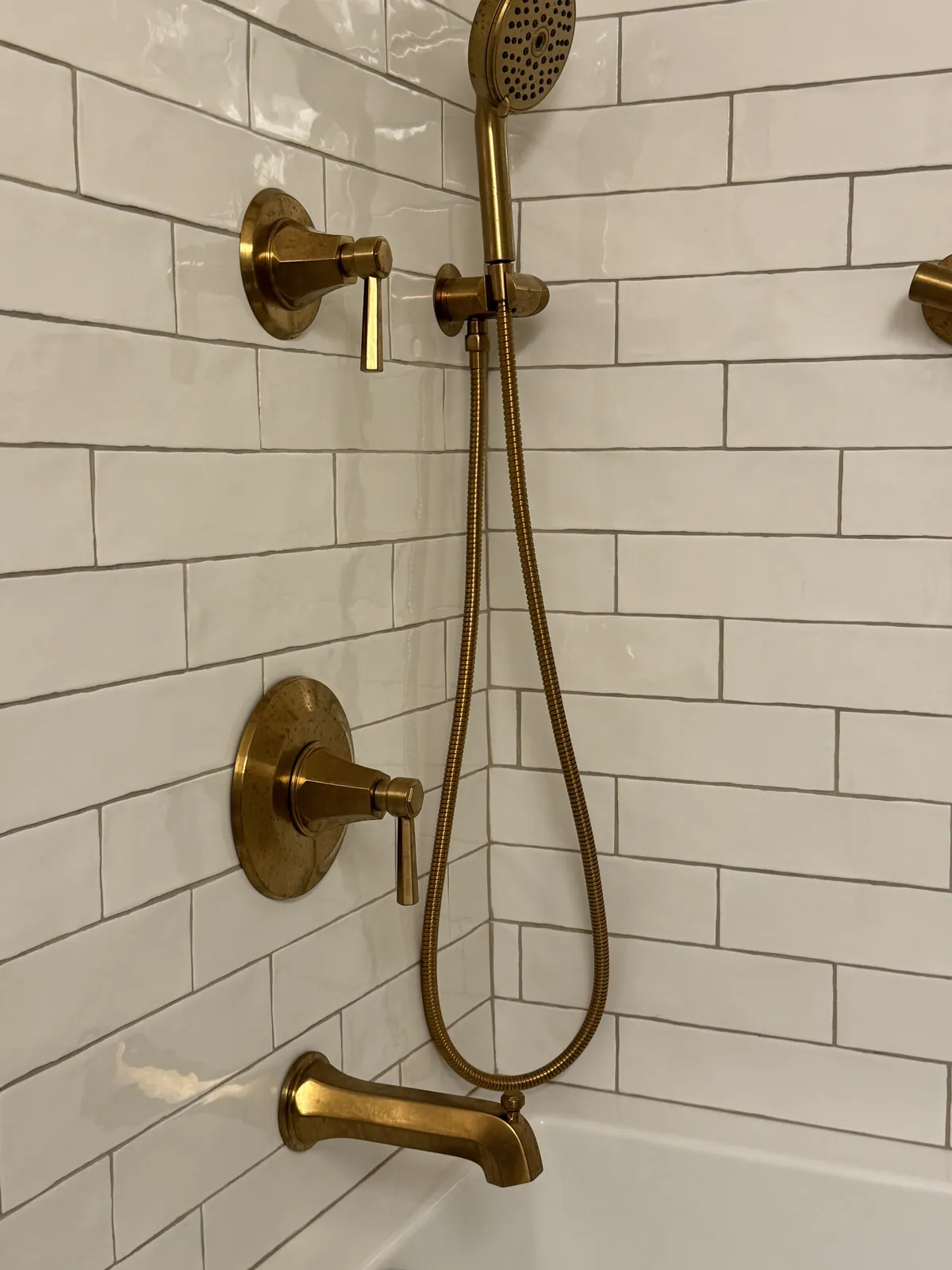Close-up of a brushed brass shower valve, hand shower and tub spout set into glossy white subway tile with gray grout