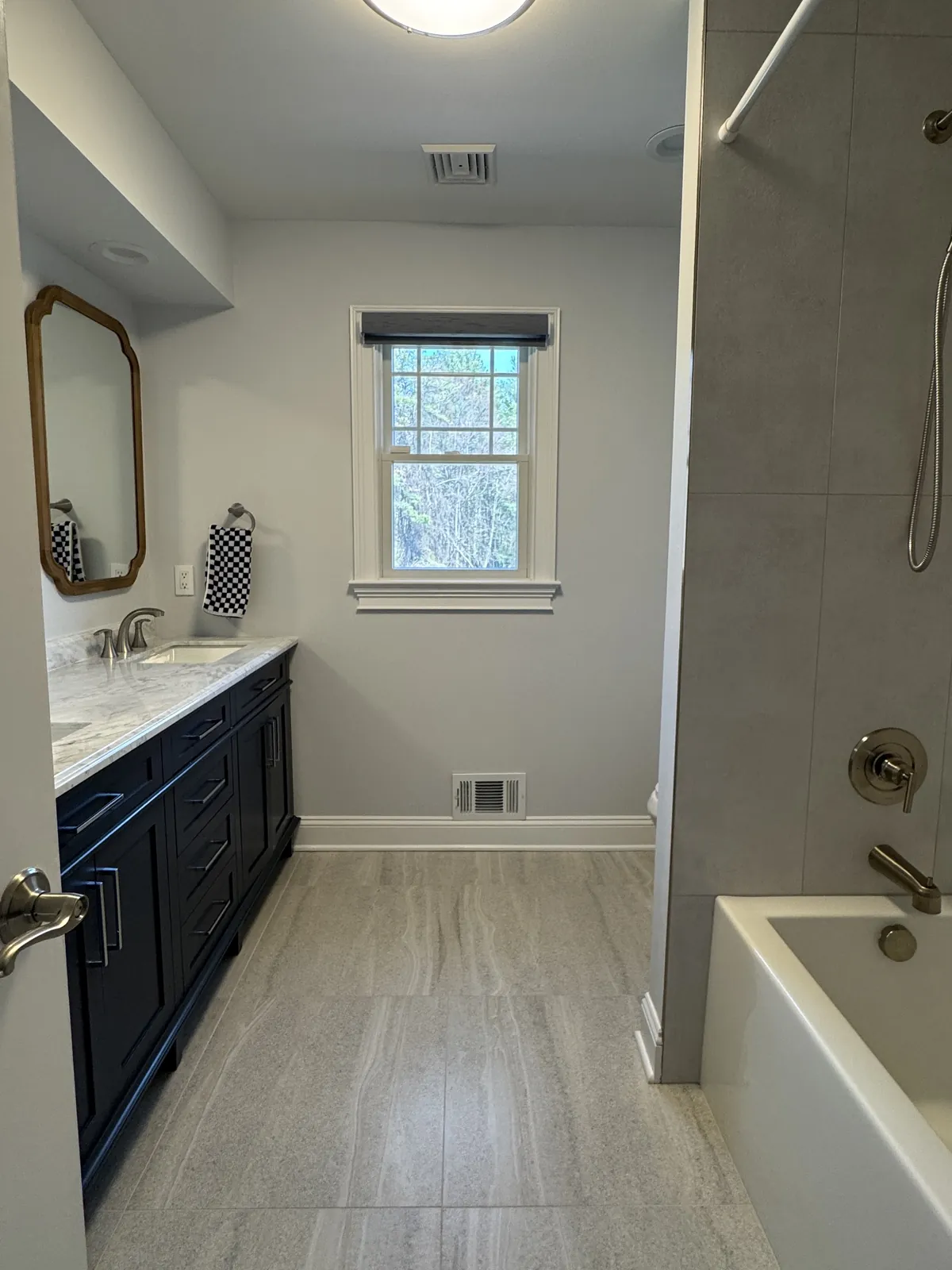 Long bathroom with a navy double vanity and marble top, brass-framed mirrors and a large-format gray floor tile running to the window