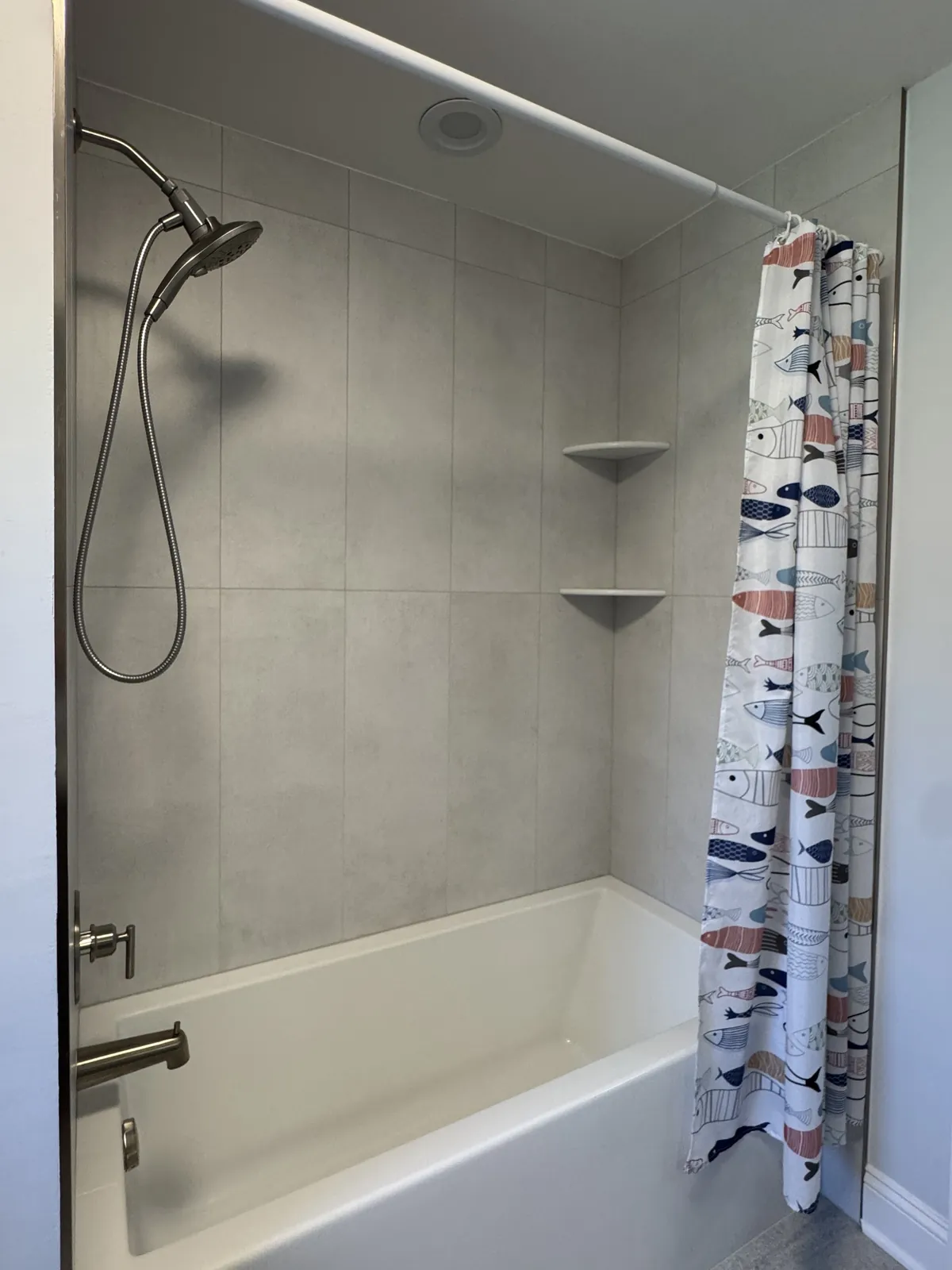 Tub surround in large-format light gray wall tile with corner shelves and a brushed nickel hand shower