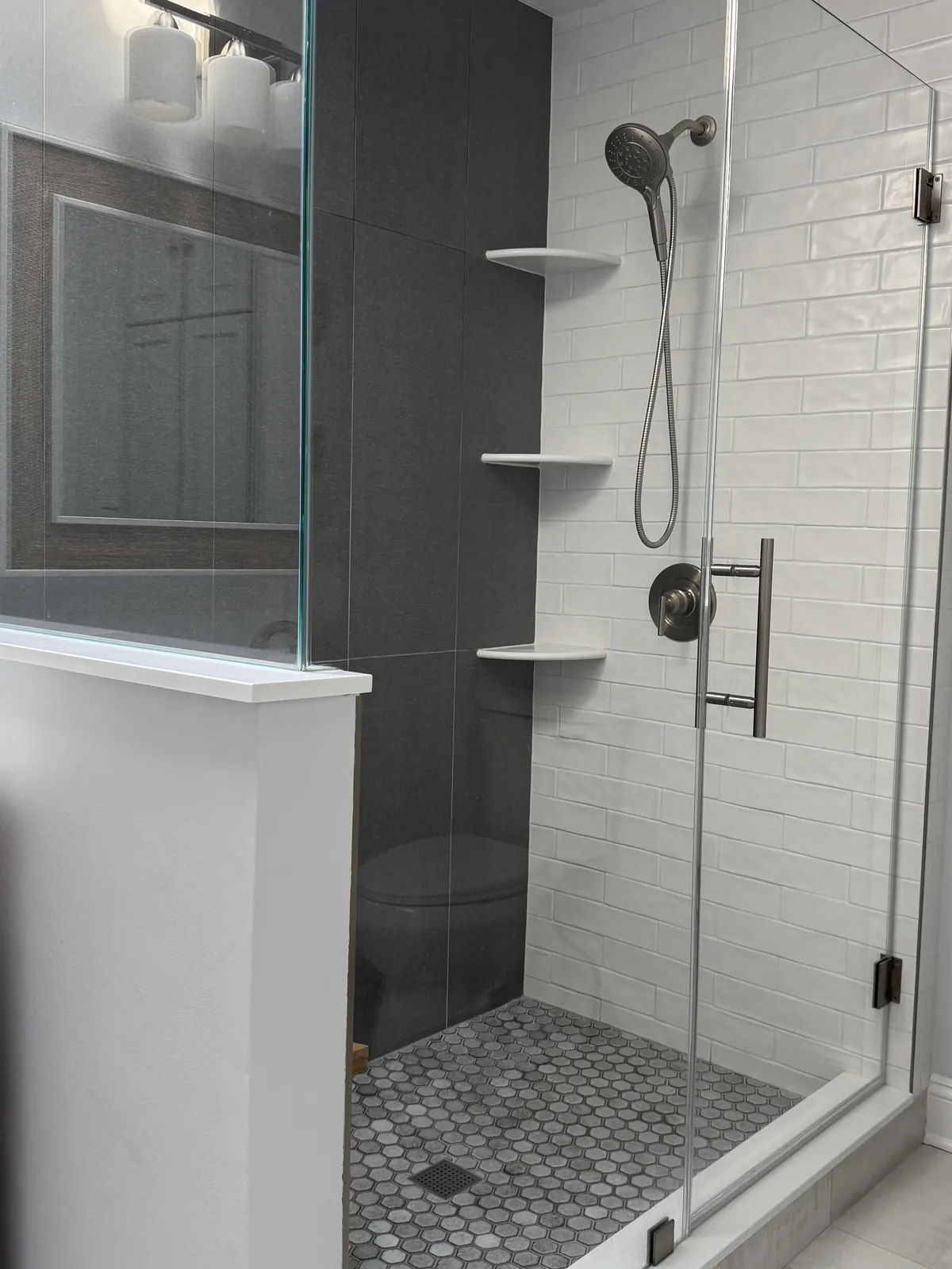 Walk-in shower behind a glass panel, white subway tile with a charcoal accent wall, corner shelves and a gray hexagon mosaic floor