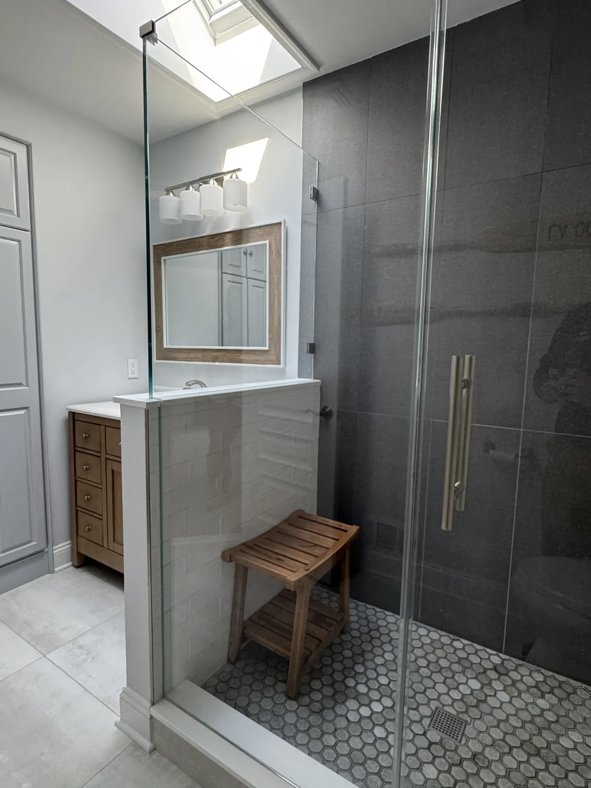 Glass-enclosed walk-in shower under a skylight, charcoal tile wall, hexagon mosaic floor and a teak bench