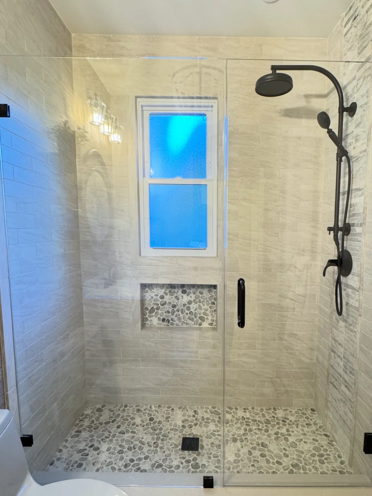 Walk-in shower seen straight on, frameless glass door, a window centred in the back wall above a pebble-lined niche, pebble floor and matte black rain shower