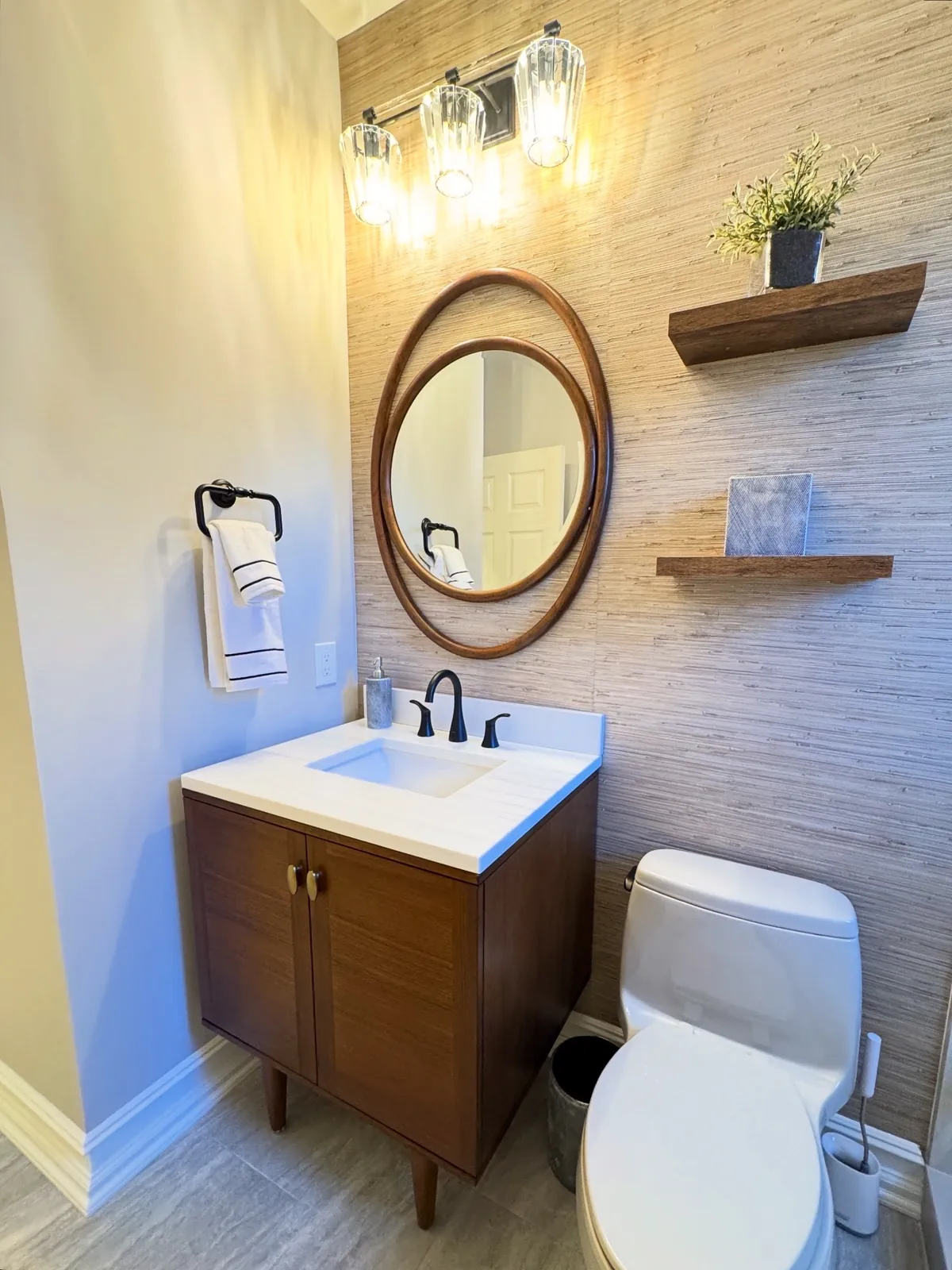 Walnut vanity under a round wood mirror on a full-height textured tile wall with floating shelves