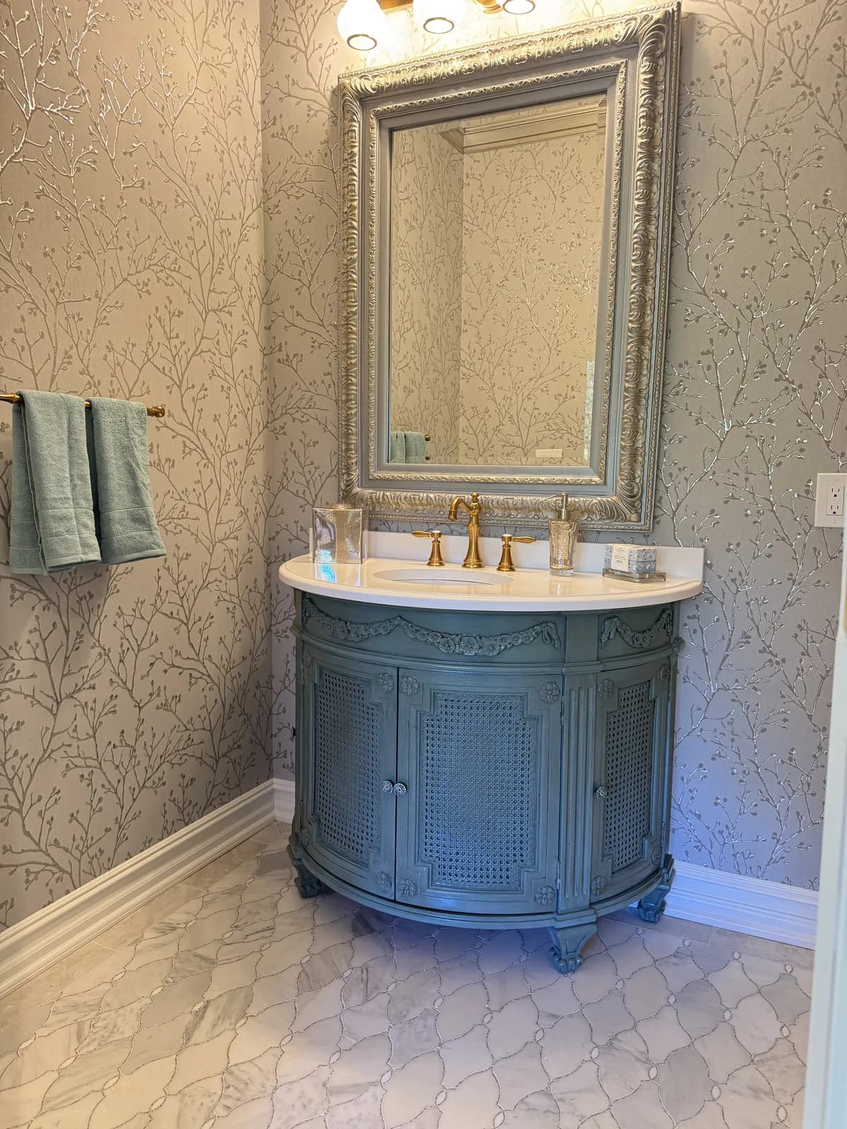 Powder room with branch-pattern wallpaper, a teal cane-front vanity with brass faucets, a silver framed mirror and a marble arabesque tile floor