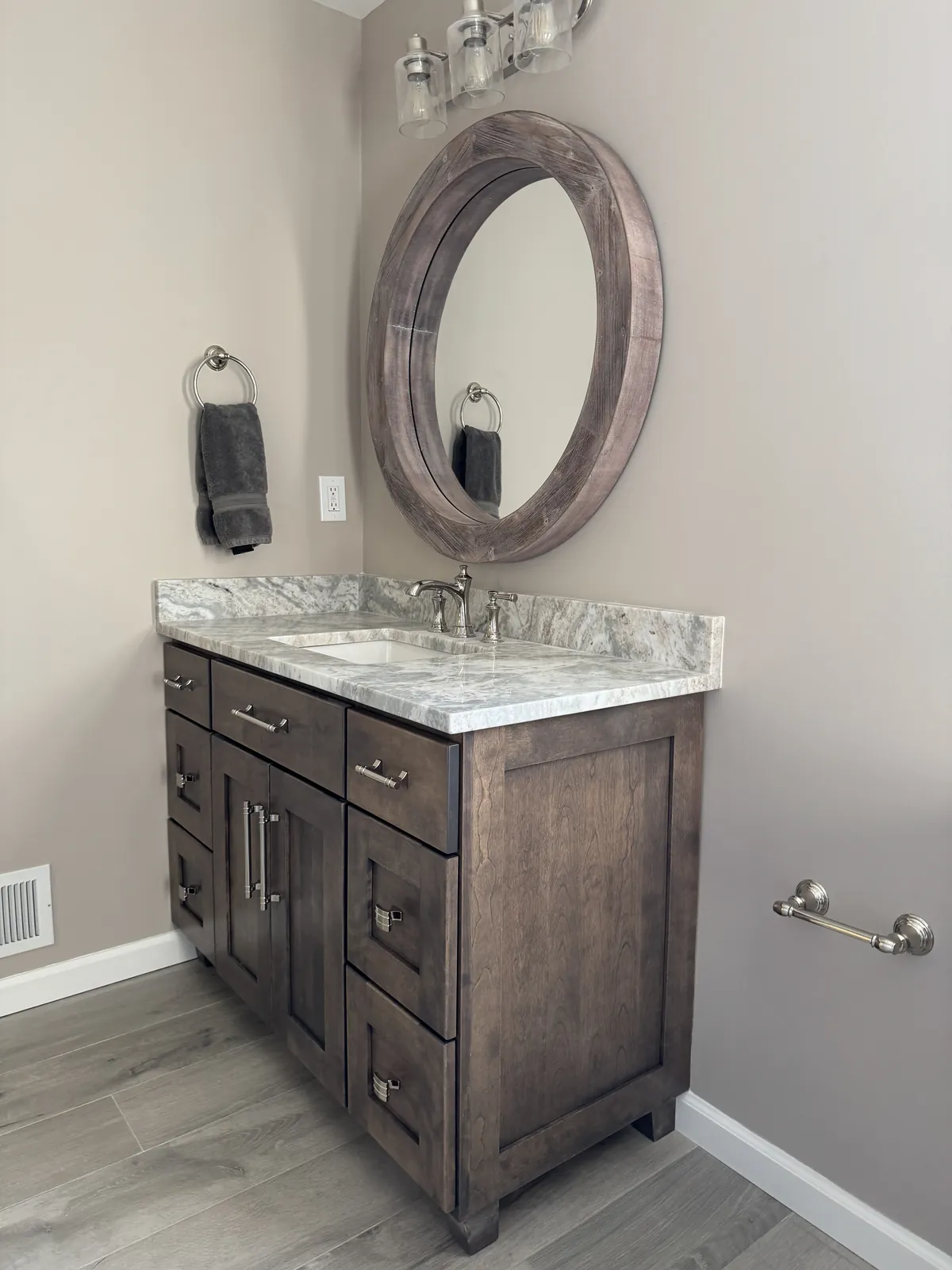 Dark wood vanity with a marble-look top under a round wood mirror, on a wood-look porcelain tile floor