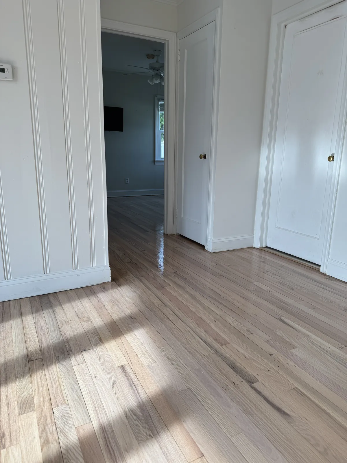 Pale oak floor with bright window light and shadow patterns, seen past a paneled doorway