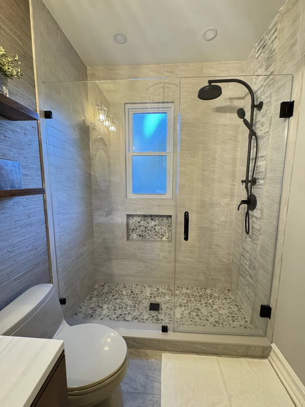 Walk-in shower with textured tile, a pebble floor and niche under the window and a frameless glass door, by Summit Home Services